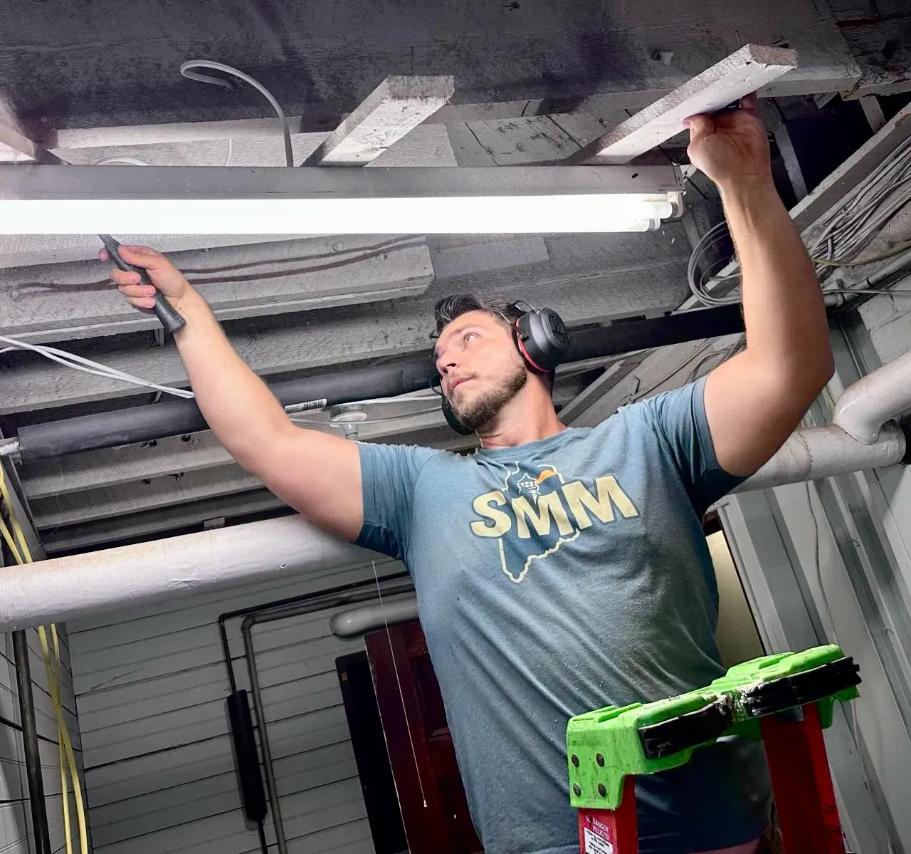 Southern Maine Mechanical technician in an SMM shirt working overhead on ceiling pipework during an HVAC job in Southern Maine
