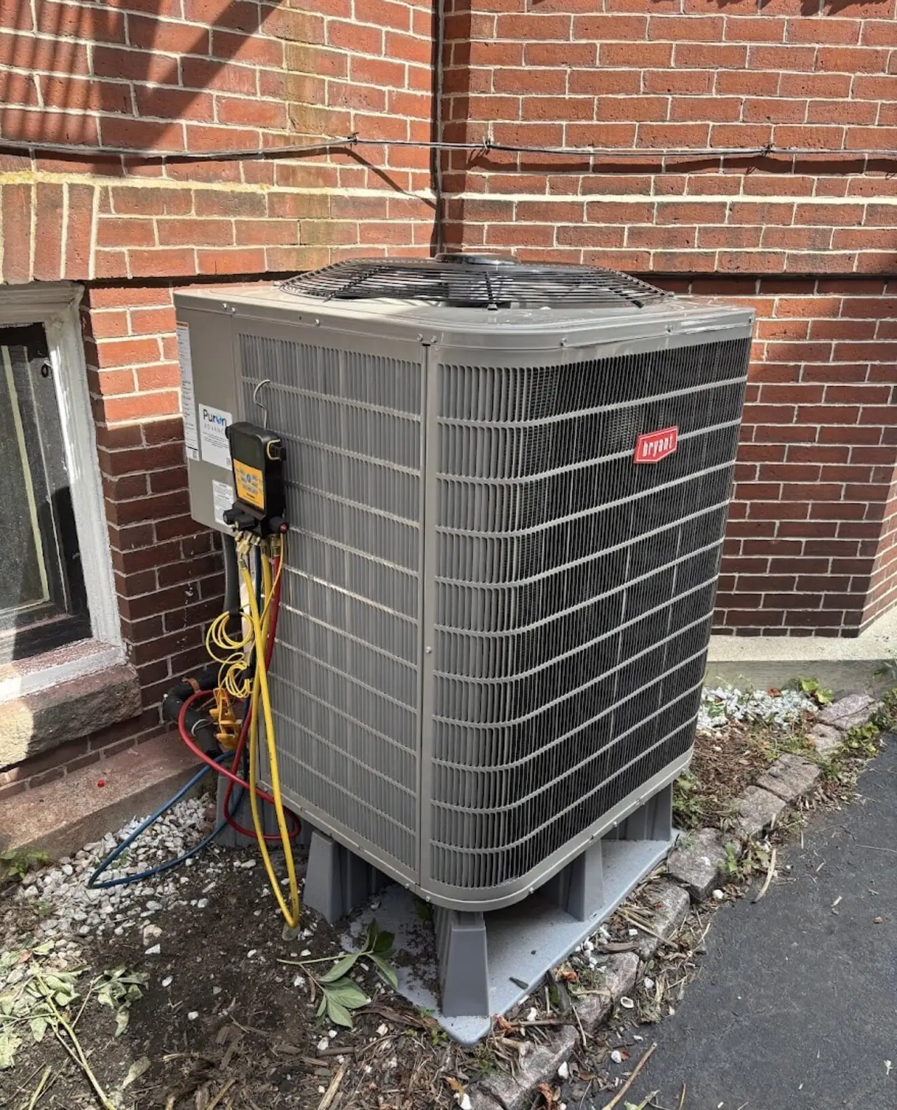 High-efficiency Bryant central air conditioning condenser installed by Southern Maine Mechanical against a brick Southern Maine home